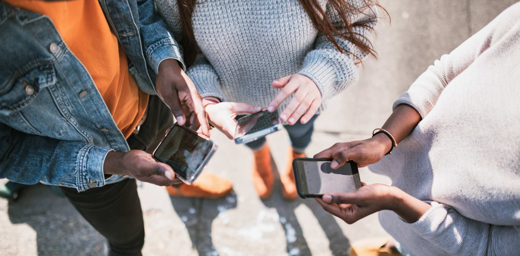 Three people stand close together outdoors, seen from above, each holding and using a smartphone. They wear casual clothes: a grey sweater, a denim jacket, and an orange shirt. Their faces are not visible, only their hands and torsos. The background appears blurred.