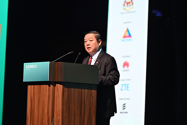 A man in a suit speaks at a wooden podium labeled “M360” on stage. Behind him is a dark background with a vertical sign displaying several company logos, including Maxis, Huawei, ZTE, and the Malaysian government crest.