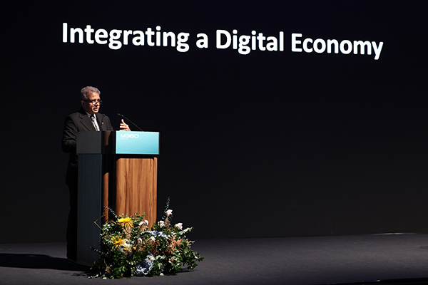 A man in a suit speaks at a podium with a “M360” sign and floral arrangement. Behind him, large text reads “Integrating a Digital Economy” against a black background. The stage is dimly lit, focusing attention on the speaker and podium.