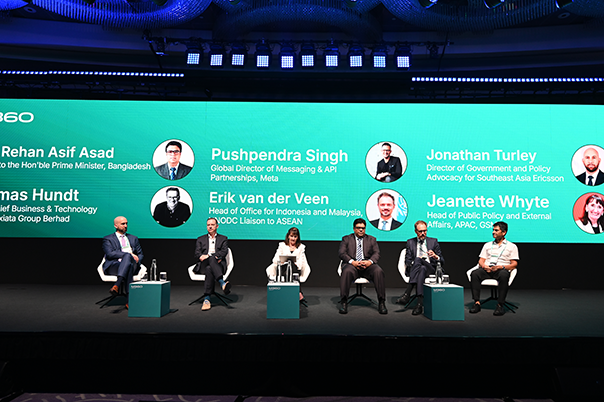 Six panelists sit on stage before a large green screen displaying their names, titles, and companies. They wear business attire, and each has a microphone. The background features circular light patterns and a modern conference setting.