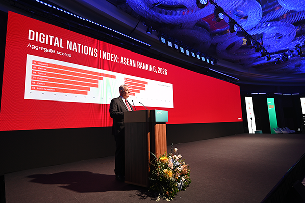 A man in a suit speaks at a podium on stage. Behind him is a large red screen displaying the “DIGITAL NATIONS INDEX: ASEAN RANKING, 2026” with a bar chart ranking countries by aggregate scores. Blue lights illuminate the ornate ceiling above.