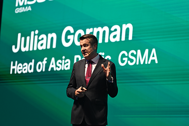 A man in a suit speaks on stage in front of a large green screen displaying the text “Julian Gorman, Head of Asia Pacific GSMA.” He gestures with one hand and holds a small device in the other. The background is brightly lit and professional.
