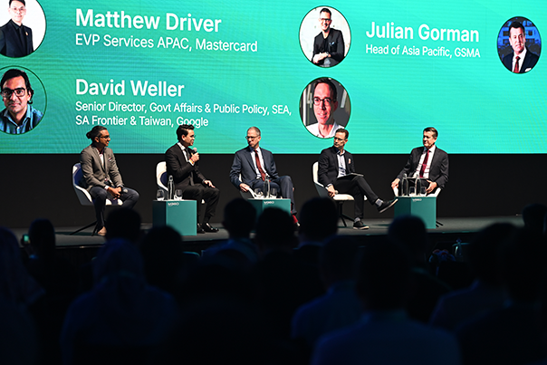 Five men in business attire sit on a stage panel, each with a microphone. Behind them, a bright screen displays their names, roles, and company photos. The audience is visible in the foreground, silhouetted against the stage lighting.