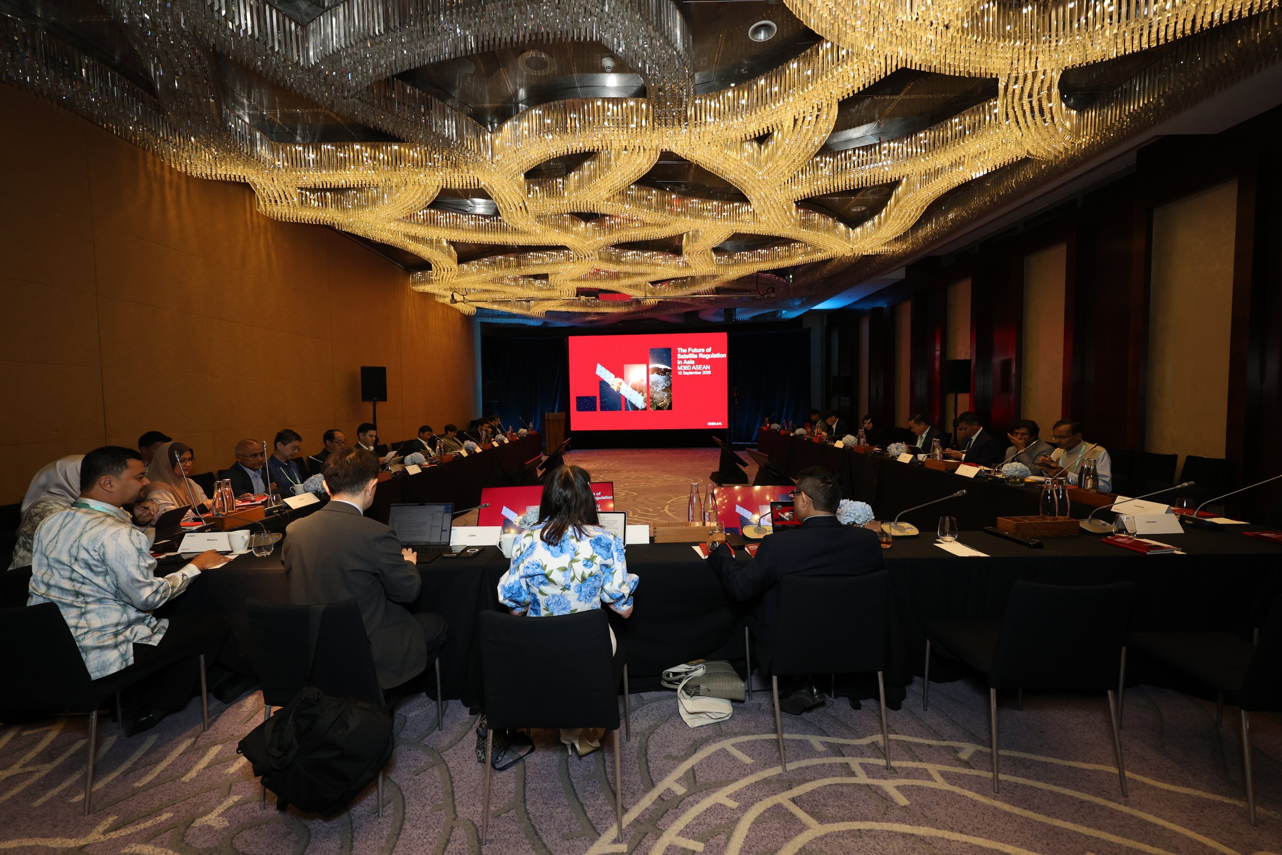 A large conference room with people seated around a U-shaped table, facing a presentation screen displaying red slides on satellite policy. The ceiling has a modern, wavy light fixture. Attendees have laptops and notebooks; the atmosphere appears formal and professional.