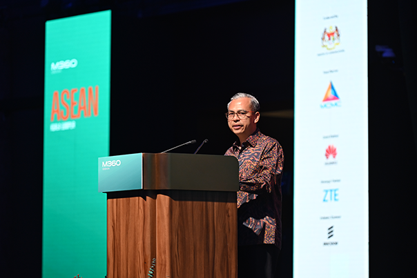 A man in glasses and batik shirt speaks at a wooden podium with “M360” and “ASEAN” on a screen. Sponsor logos, including Huawei and ZTE, are visible on a tall vertical display beside him. The background is dark, highlighting the speaker.