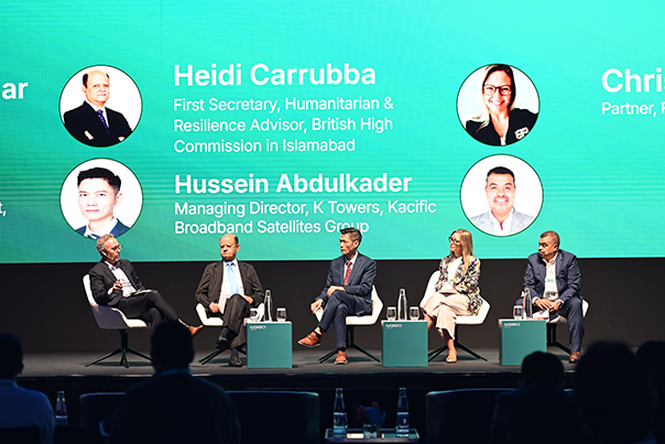 Five panelists sit onstage in chairs, speaking at a conference. Behind them is a green screen displaying names, titles, and photos, including Heidi Carrubba and Hussein Abdulkader. Water bottles and microphones are on small tables in front of the panelists.