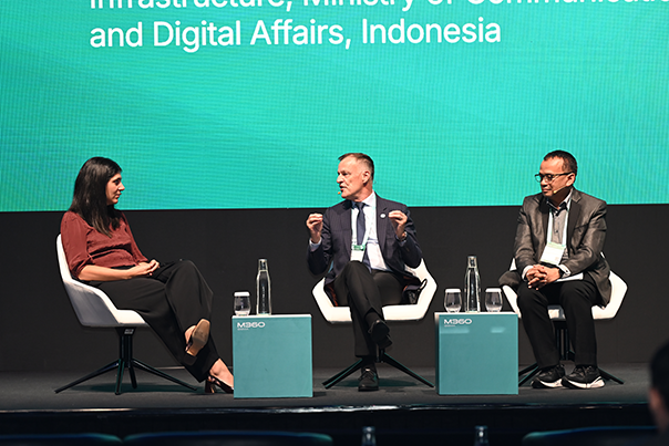 Three people sit on stage in white chairs, having a panel discussion. Two men in suits and a woman in a maroon shirt face each other. Behind them, a green screen displays text mentioning “Ministry of Communications and Digital Affairs, Indonesia.”.