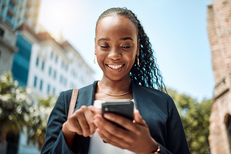 A woman with long, curly hair smiles while using a smartphone outdoors, perhaps checking the latest updates in mobile industry public policy. She wears a navy blazer, gold necklace, and carries a brown shoulder bag. Tall buildings and green trees are blurred in the sunny background.