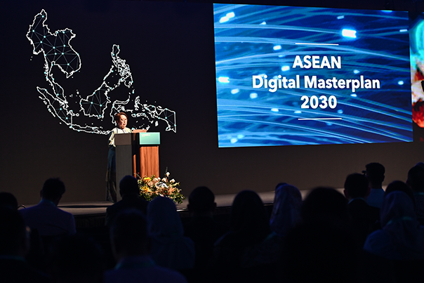 A speaker stands at a podium on stage beside a large screen displaying “ASEAN Digital Masterplan 2030.” An illuminated map of Southeast Asia is visible in the background. The audience sits in shadow in the foreground.
