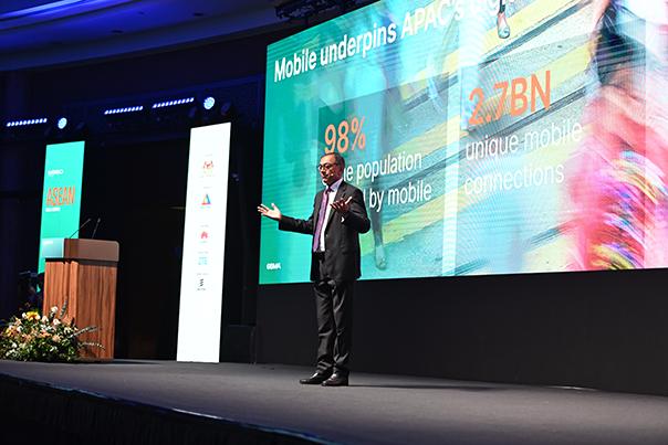 A man in a suit gives a presentation on stage beside a large screen showing mobile usage stats: “98% of the population covered by mobile” and “2.7BN unique mobile connections.” The room is dimly lit, with spotlights and a flower arrangement on stage.