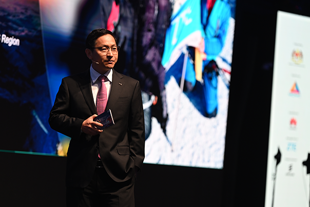 A man in a dark suit, pink tie, and glasses stands on stage holding a phone. Behind him is a large blurred screen with people and bright colors. Logos are visible on a vertical white panel to the right. The stage is dimly lit.