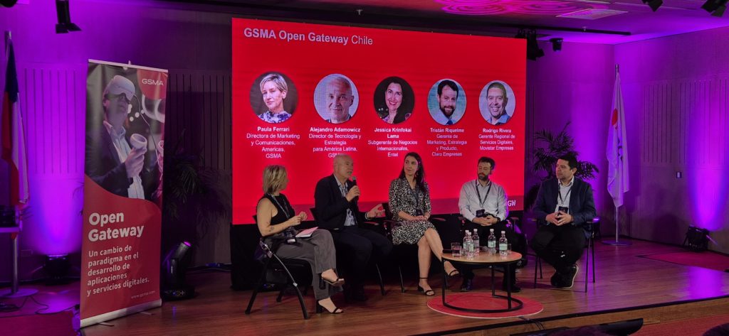 A panel of five speakers sits on stage at the GSMA Open Gateway Chile event. Behind them is a red screen displaying their names and roles. A banner to the left highlights the event. The room is softly lit with purple lighting, and a small audience is visible in front.