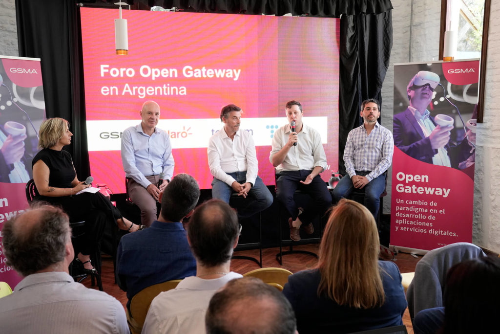 A panel of five people, four men and one woman, are seated on a stage at the "Foro Open Gateway en Argentina." They face an audience while discussing topics. Behind them are red banners with the event logo and text promoting digital applications and services. The room has a casual, professional setting.