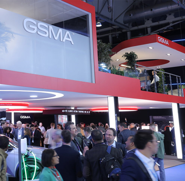 A crowded tech event buzzes with energy as people mingle around GSMA's modern exhibition booth. Featuring two levels, the upper deck boasts plants and a lounge area perfect for discussions on network APIs. Bright lights and a high ceiling enhance the bustling atmosphere.