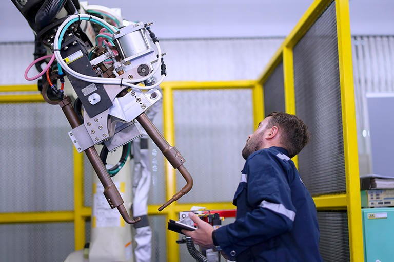A technician in a blue work jacket operates a large industrial robotic arm, integrated with network APIs, inside a factory. The arm is equipped with various cables and metallic components. The technician holds a remote control device, focusing on the machine, with a safety enclosure visible in the background.