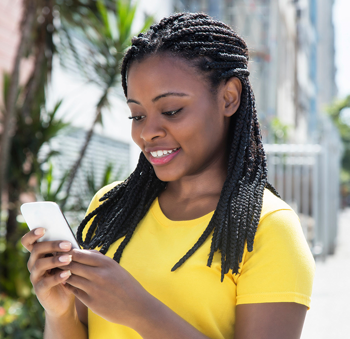 A woman with braided hair, wearing a yellow shirt, smiles while looking at her smartphone. She is standing outdoors on a sunny day, with a blurred background of trees and buildings.