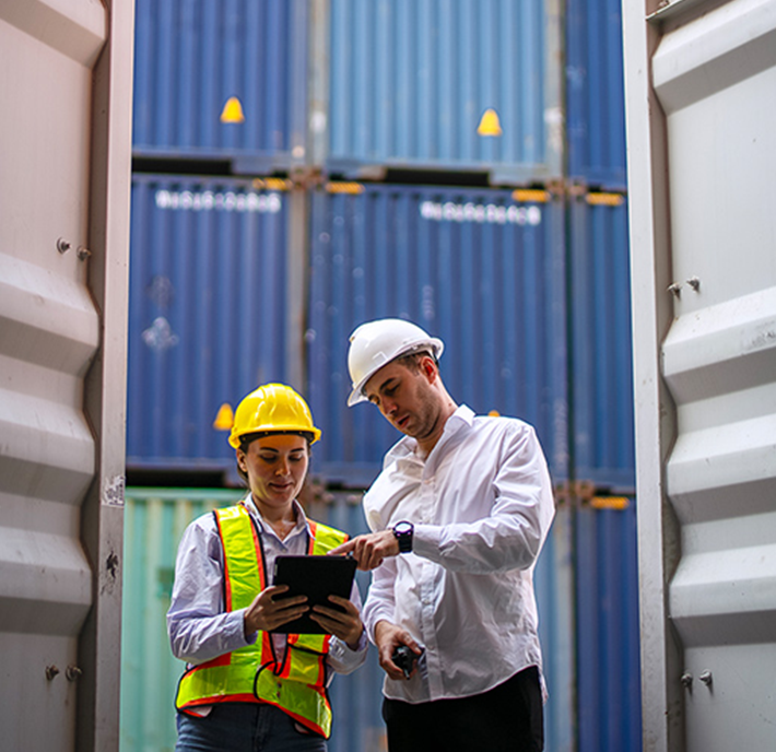 Two people stand between shipping containers. One wears a yellow hard hat and reflective vest, holding a tablet displaying network APIs. The other, in a white hard hat, points at the tablet. Stacked blue containers are visible in the background as they discuss logistics.