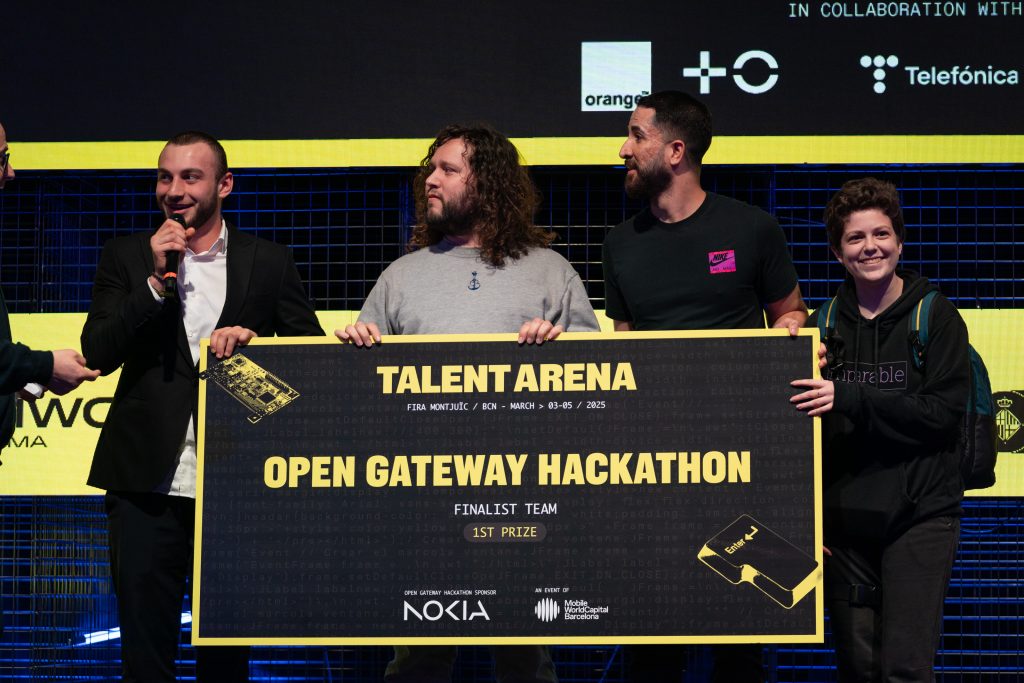 Four people on stage smiling, holding a large check for "Open Gateway Hackathon Finalist Team 1st Prize" at the Talent Arena event. The background features logos and a yellow and black color scheme. The individuals are casually dressed and appear celebratory.