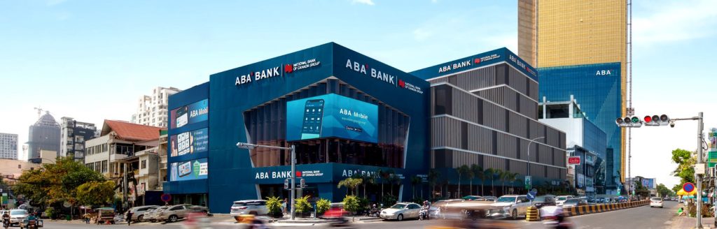 A busy city intersection with a large, modern ABA Bank building featuring blue signage and glass windows. Motorbikes and cars pass by on the street, and tall office buildings rise in the background under a clear blue sky.