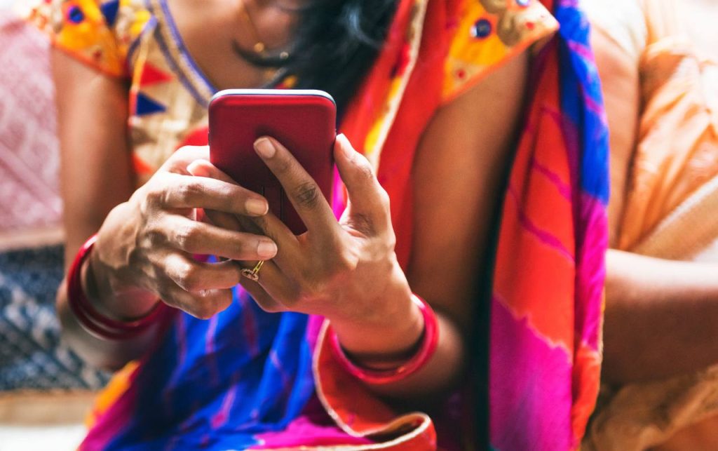 A woman in a colorful saree, wearing red bangles and a ring, is holding and using a red smartphone. The focus is on her hands and phone; her face is not visible. The bright saree features orange, blue, and red patterns.