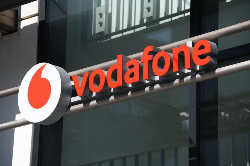 The image shows a Vodafone store sign with the red Vodafone logo next to the word “vodafone” in bold red letters on a modern glass building exterior, with some metal bars and reflections visible in the background.