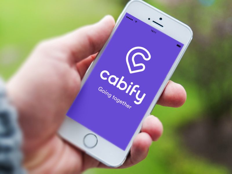 A hand holds a white smartphone displaying the Cabify app’s purple loading screen, featuring the Cabify logo and tagline “Going together.” The background is blurred, showing green foliage and suggesting an outdoor setting.