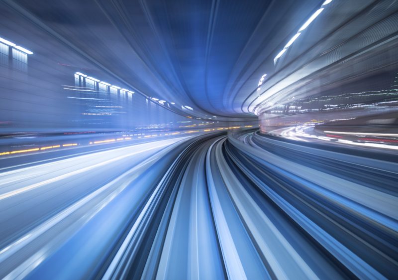 A long-exposure photo captures blurred blue and white light trails along a curving train track in an urban tunnel at night, creating a sense of high speed and motion. The lights streak both horizontally and vertically.