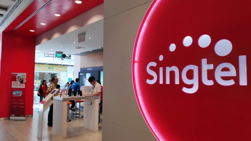 A Singtel store interior with a large, illuminated red and white Singtel logo on the wall, counters with electronic devices, and people interacting with staff. The setting appears modern and bright, with a mall visible in the background.