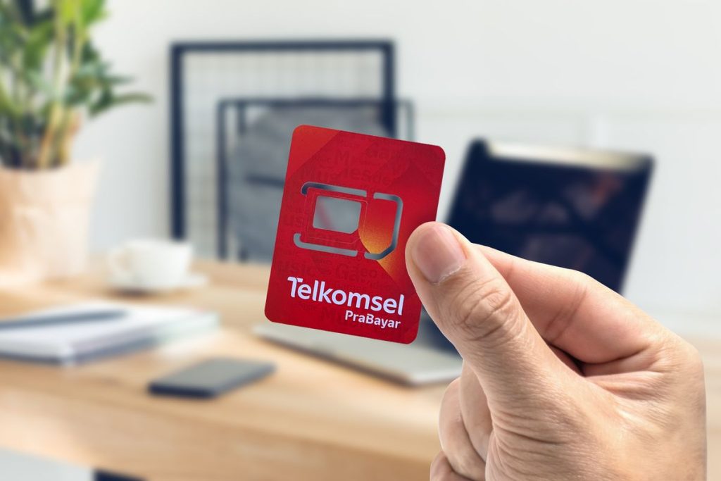 A hand holds a red Telkomsel Prabayar SIM card in focus. In the blurred background, there is a wooden desk with a laptop, tablet, papers, and office supplies, suggesting a work or home office setting.