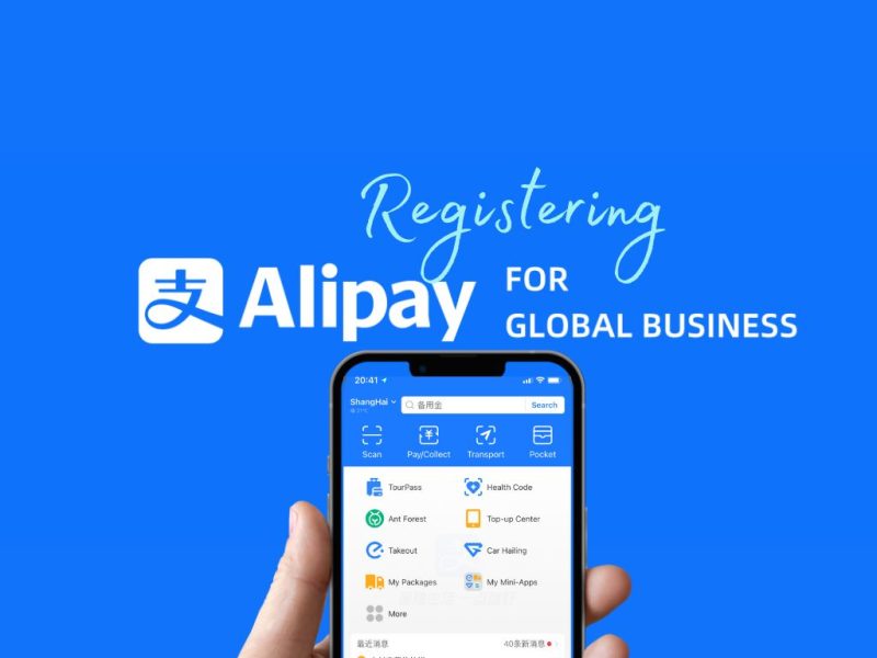 A hand holds a smartphone displaying the Alipay app interface. The blue background features the Alipay logo and text: “Registering Alipay for Global Business.” The app screen shows menu icons for services like Scan, Pay/Collect, Transport, and Pocket.