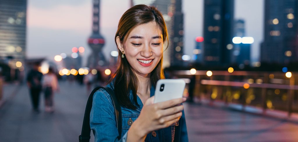 A young woman with long brown hair smiles while looking at her smartphone, connected via a 5G network-as-a-service NaaS API open-gateway. She stands outdoors in a city at dusk, with blurred tall buildings and bright lights behind her.