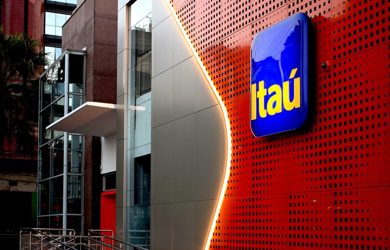 A modern building exterior with a perforated red metal wall curves around a gray panel. A blue Itaú bank logo is illuminated on the wall, with soft yellow accent lighting emphasizing the curve. Glass and steel architectural elements are visible on the left side.