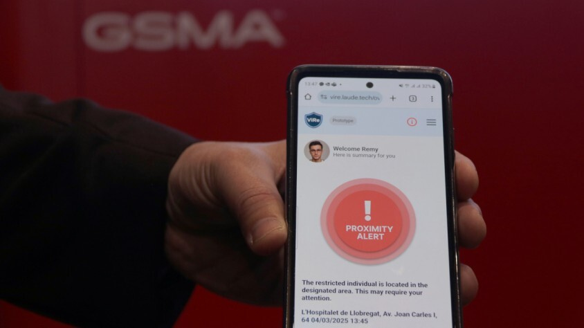 A hand holds a smartphone displaying a "PROXIMITY ALERT" screen with a red circular warning icon. The alert mentions a restricted individual's location. The background is blurred red with the letters "GSMA" partially visible.