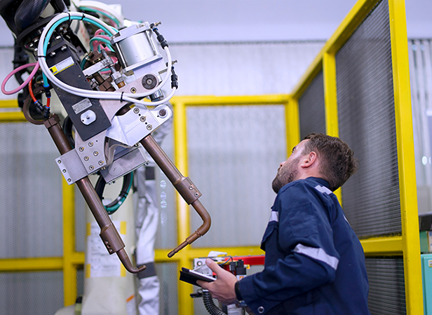 A man in industrial workwear operates and inspects a large robotic arm in a factory setting. He holds a control device while looking up at the machinery. Yellow safety barriers and metal fencing surround the workspace.