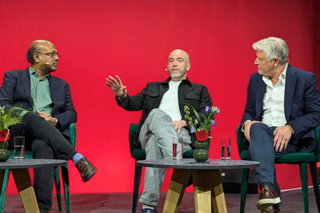 Three men in business attire sit on green chairs against a bright red background, engaged in conversation. The man in the center gestures while speaking, as the other two listen. Two small tables with flowers and glasses of water are in front of them.