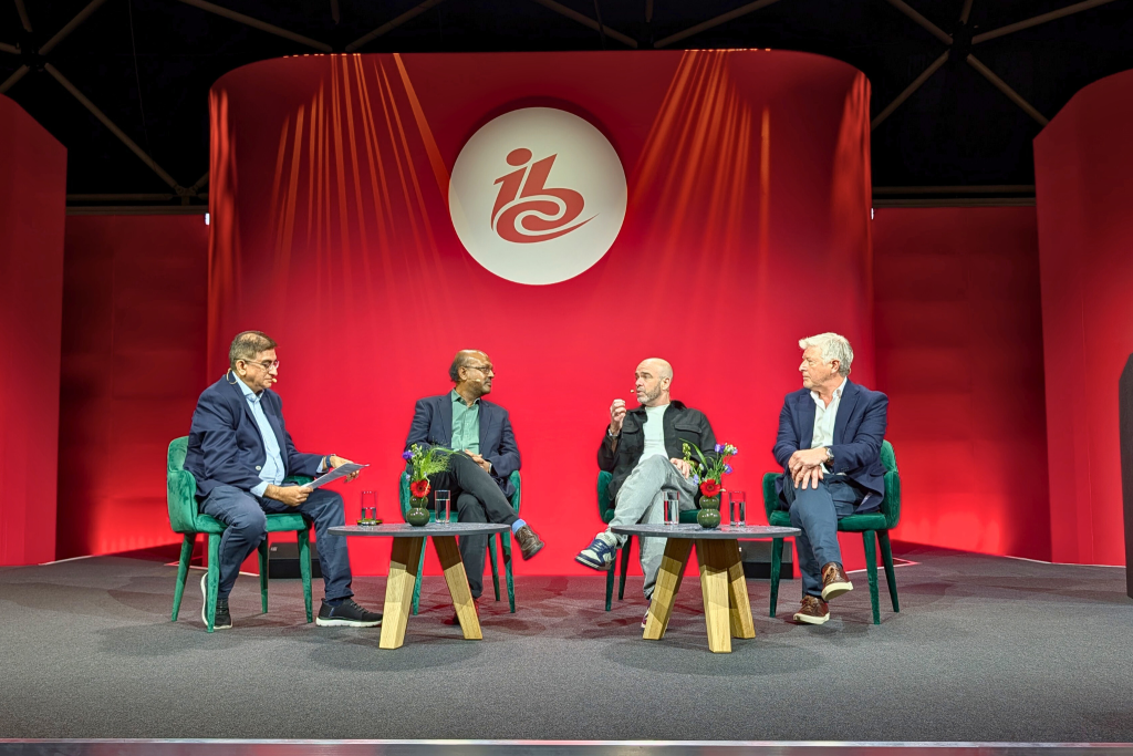 Four men sit on stage in armchairs, engaged in a panel discussion. They are surrounded by small tables holding water glasses and flowers. Behind them is a red backdrop with the IBC logo in white. The atmosphere is professional and focused.