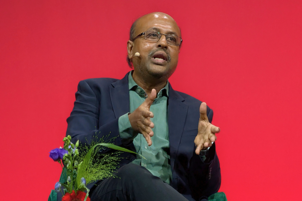 A man with glasses, a bald head, and a short beard speaks while gesturing with his hands. He wears a dark blazer and green shirt, has a headset microphone, and sits in front of a red background. A bouquet of flowers is visible in the lower left corner.