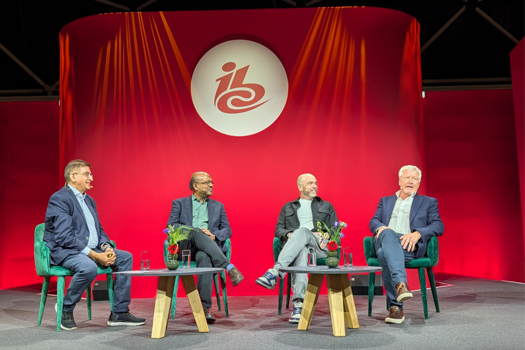 Four men sit onstage in armchairs, engaged in a panel discussion. They wear business-casual clothing, with glasses of water and small vases of flowers on tables before them. The backdrop is bright red with a white circular logo containing the letters “ibc.”.