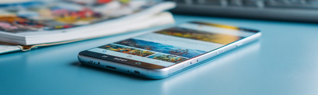 A smartphone lies face up on a blue desk, displaying a gallery of colorful photos. Next to it is an open magazine with blurred images. The background includes part of a keyboard, suggesting a workspace or creative environment.