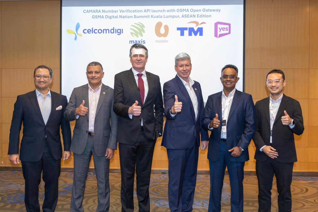 Six men in business attire stand side by side, smiling and giving a thumbs-up. Behind them is a screen displaying logos for CelcomDigi, Maxis, U Mobile, TM, and Yes, and text referencing the GSMA Digital Nation Summit in Kuala Lumpur, ASEAN Edition.