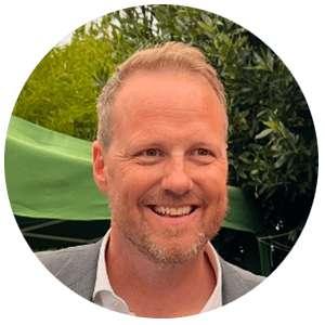 A smiling man with short blond hair and a light beard is wearing a gray suit jacket over a white shirt. He is outdoors, with green leafy trees and a green tent canopy visible—perhaps at an event focused on Aviation APIs. The photo is cropped in a circular frame.