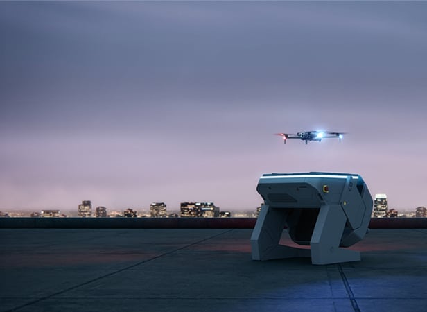 A small Skydio drone with red and blue lights hovers above a rooftop at dusk. In the foreground, a futuristic rectangular device sits on the rooftop, with the illuminated city skyline visible under a cloudy sky.