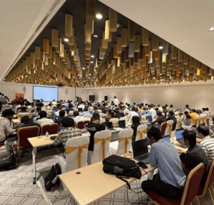 A large conference room is filled with people seated at rows of tables, many using laptops. The ceiling features gold and brown cylindrical decorations. A projector screen and speakers are visible at the front, and the room is well-lit and modern.