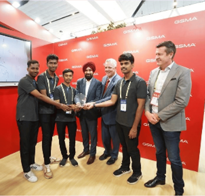 Five young men in black shirts accept a trophy from two men in suits, standing on a wooden floor with a red GSMA backdrop. A television screen is mounted on the left wall. All subjects are smiling and looking at the camera.