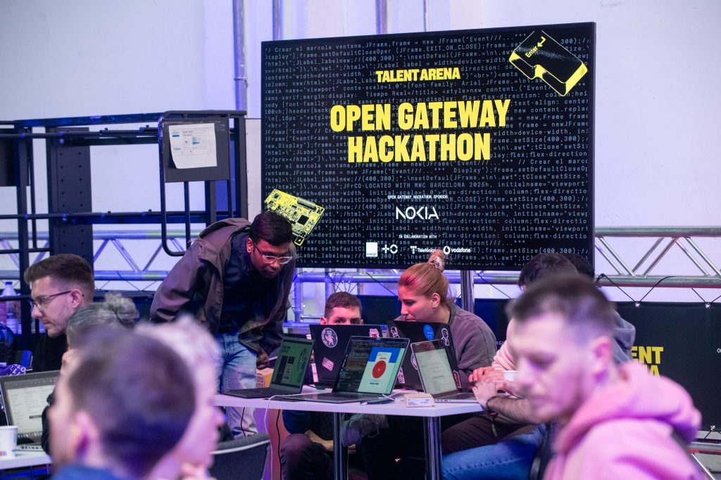 A group of people work on laptops at round tables during a hackathon. Behind them, a large screen displays “Talent Arena Open Gateway Hackathon” with logos and a tech-themed background. The atmosphere is collaborative and focused.