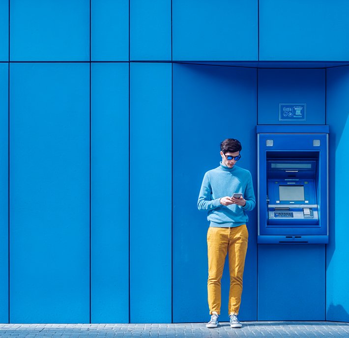 A man in a blue sweater and yellow pants stands by a modern blue ATM, possibly accessing financial services APIs on his phone. The colorful scene features geometric wall panels and soft sunlight, creating a vibrant, contemporary atmosphere.