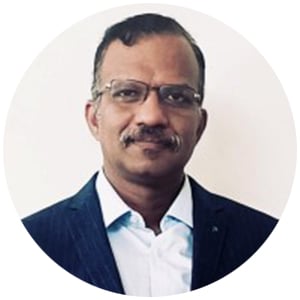 A middle-aged man with short dark hair, glasses, and a mustache, wearing a dark striped suit jacket over a light blue shirt, stands against a plain white background. He faces forward with a neutral expression. The circular image evokes Tata Elxsi's professionalism.