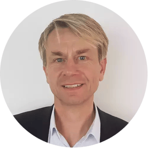A middle-aged man with light skin, short blond hair parted to the side, and blue eyes is smiling slightly. He is wearing a black blazer over a light blue collared shirt—classic professional style for a FICO executive. The photo is cropped in a circular frame.