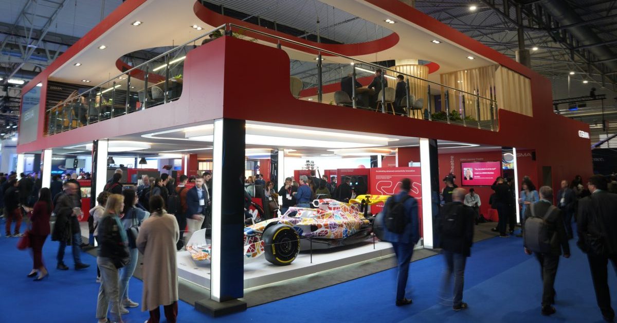 A colourful Formula 1 racing car is displayed under a modern, two-storey red GSMA stand at a busy indoor exhibition. People walk around the car and gather on both exhibition floors; blue carpet covers the venue floor, and large ceiling lights illuminate the scene.