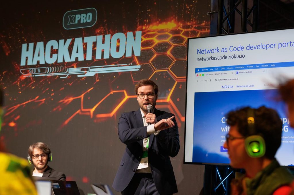 A man speaks on stage at a hackathon event, pointing, with a large screen and logo behind him.